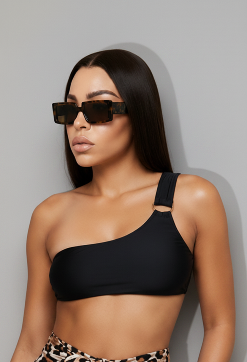 Model wearing a black one-shoulder bikini top paired. The look is completed with rectangular tortoise sunglasses against a light gray studio background.