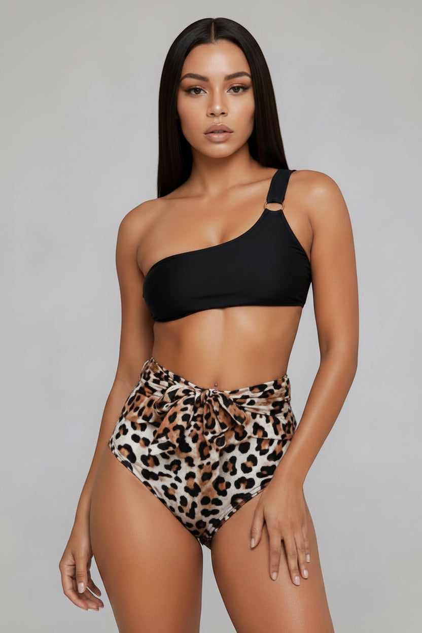 A woman modeling a two-piece swimsuit with a black one-shoulder bikini top and high-waisted leopard print bikini bottoms, set against a plain light gray studio background.